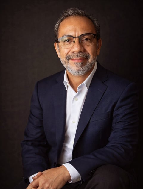 Professional headshot of a middle-aged man wearing glasses, dark blazer, and white shirt against a dark background