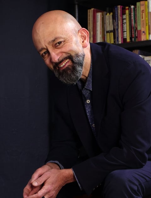 Bald man with full beard wearing dark blue blazer and patterned shirt, smiling at camera with bookshelves in background